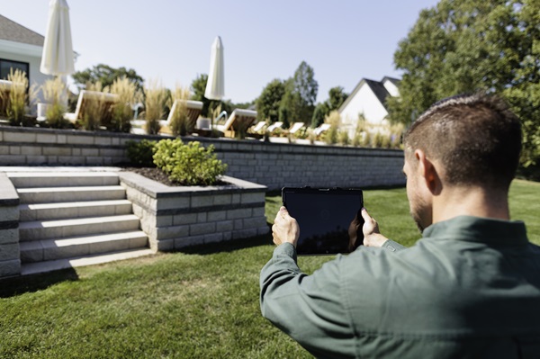 A property inspector with a green shirt holds up a tablet and views exterior stairs and retaining walls.