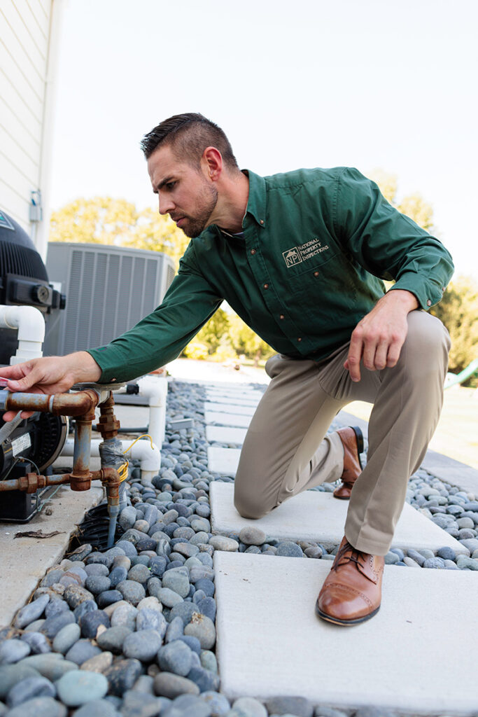 An NPI inspector examines exterior piping while kneeling outside a building.